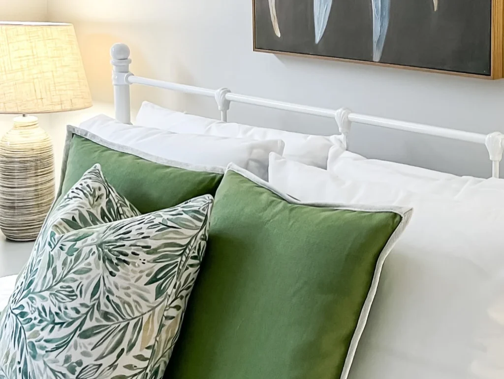 A white bed with green pillows and a painting on the wall.